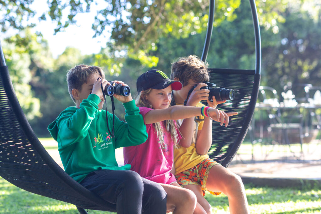 3 children on a outdoor swing with binoculars and a camera
